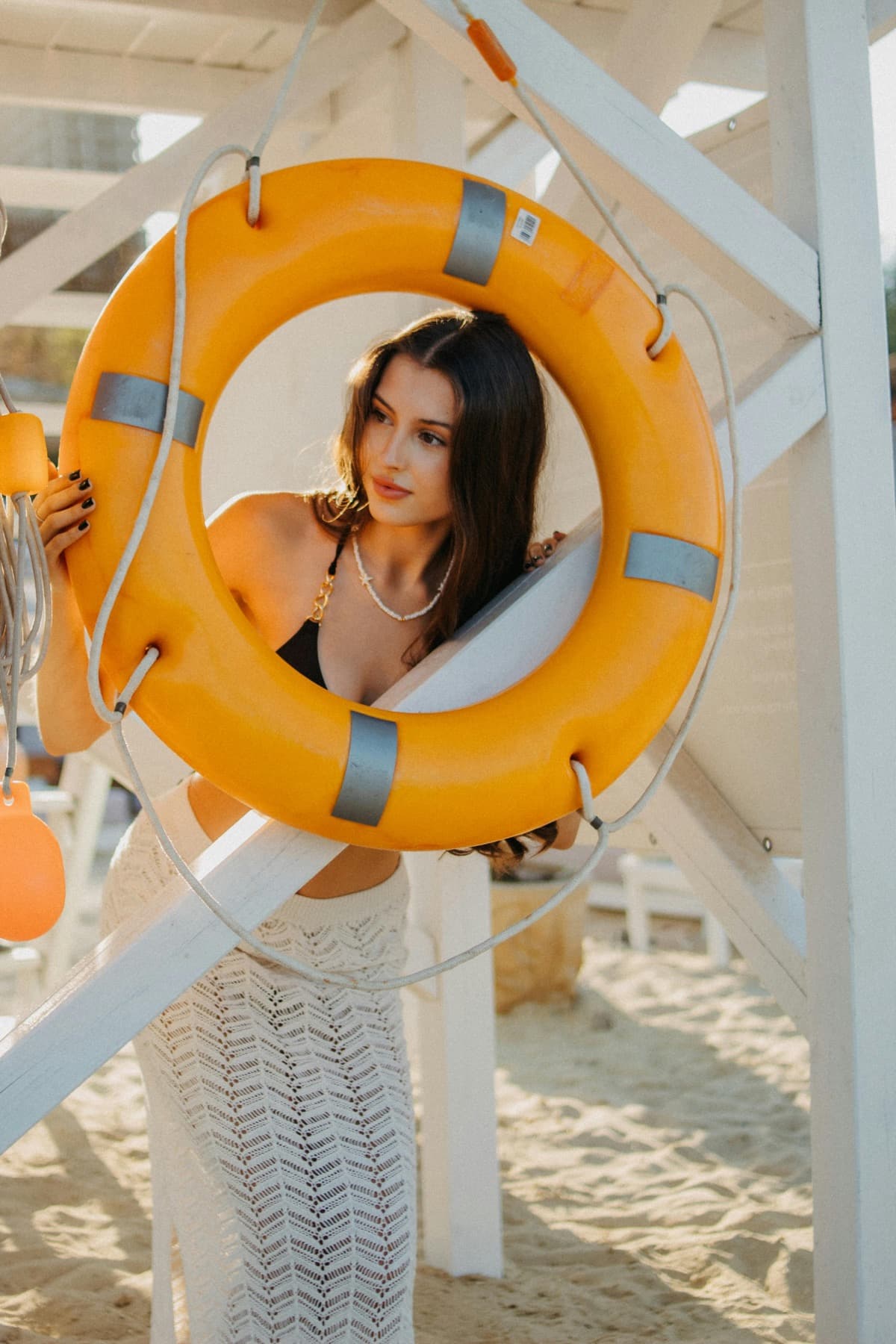 A woman on a beach looking out through an orange life ring hung on a white lifeguard tower.